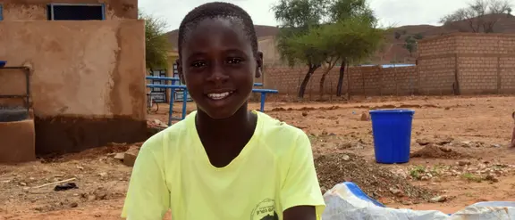 Lächelnder Junge sitzt vor einem Gebäude in einer ländlichen Gegend von Burkina Faso, umgeben von einfacher Architektur und weiter Landschaft.