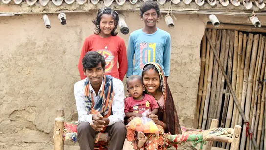 Eine indische Familie sitzt vor einem Lehmhaus, wobei die Eltern mit ihrem Kleinkind auf einer Bank sitzen und die älteren Kinder dahinter stehen.