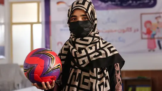Junge Frau in gemustertem Kopftuch und Maske hält einen bunten Fußball in einem Raum mit Banner im Hintergrund.
