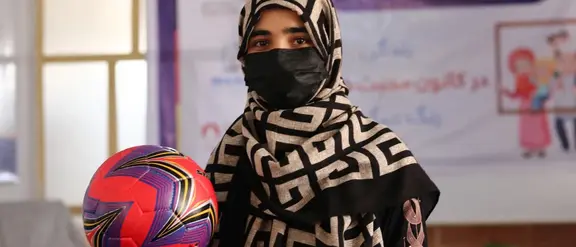 Junge Frau in gemustertem Kopftuch und Maske hält einen bunten Fußball in einem Raum mit Banner im Hintergrund.