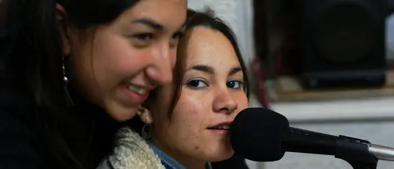 Zwei junge Frauen in einem Tonstudio, eine spricht in ein Mikrofon, die andere lächelt daneben.
