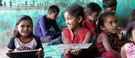 Lachende Kinder in einem Klassenzimmer in Indien sitzen auf dem Boden und halten Tafeln. Die Szene zeigt eine lebendige und kreative Lernumgebung, die die Freude am Lernen und die kulturelle Vielfalt in Indien betont.
