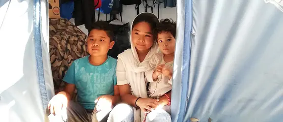 Drei Kinder sitzen im Eingang eines Zeltes in einem Flüchtlingslager in Griechenland, vereint in ihrem Zusammenhalt.