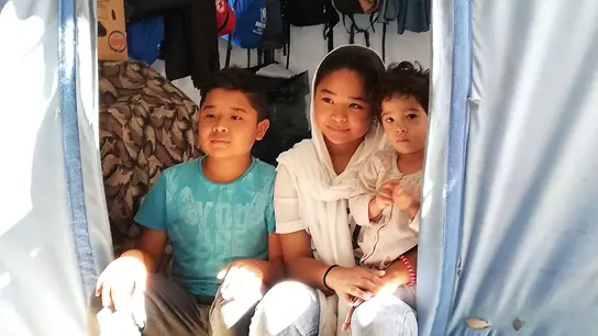 Drei Kinder sitzen im Eingang eines Zeltes in einem Flüchtlingslager in Griechenland, vereint in ihrem Zusammenhalt.
