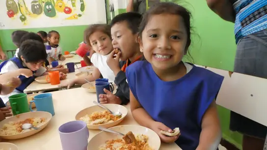  Lächelndes Mädchen sitzt mit anderen Kindern in einer Kantine in El Salvador und isst eine Mahlzeit.
