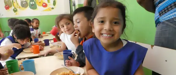  Lächelndes Mädchen sitzt mit anderen Kindern in einer Kantine in El Salvador und isst eine Mahlzeit.