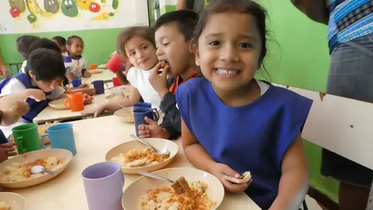  Lächelndes Mädchen sitzt mit anderen Kindern in einer Kantine in El Salvador und isst eine Mahlzeit.