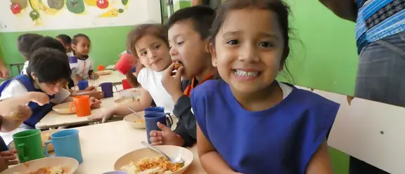  Lächelndes Mädchen sitzt mit anderen Kindern in einer Kantine in El Salvador und isst eine Mahlzeit.